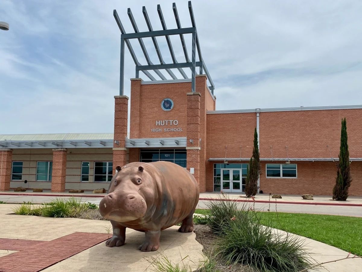 One of Hutto's signature concrete hippo statues — the city's beloved "Hippo Nation" mascot — standing in front of a Hutto ISD school building under a clear blue Texas sky