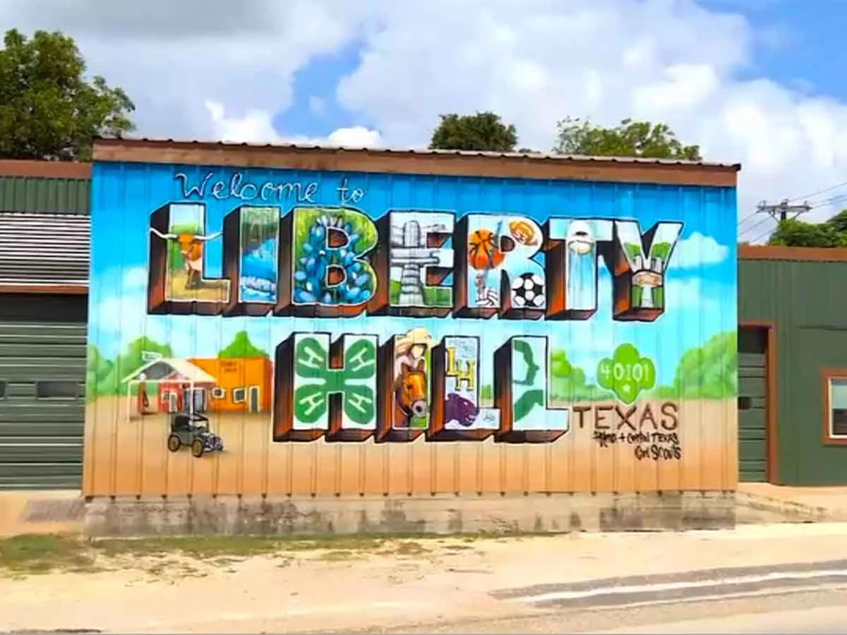 A colorful "Welcome to Liberty Hill, Texas" community mural painted on a downtown metal building — oversized block letters filled with local icons including a longhorn, bluebonnets, a windmill, the Liberty Hill Panther, sports, and 4-H clovers, under a blue Hill Country sky